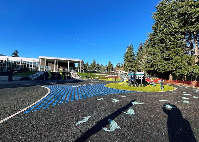 outdoor play area with a play structure, fish and filled circles painted on paving, and two slides on a slope from a building to the play area